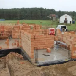 Budowa domu jednorodzinnego w trakcie wznoszenia ścian z czerwonej cegły, widoczne zbrojenia i fundamenty, w tle las i biały budynek gospodarczy.
