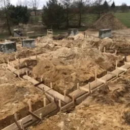 Przygotowanie fundamentów pod budowę domu: wykopane rowy z drewnianym szalunkiem, widoczne betonowe bloczki na obrzeżach terenu, w tle nasyp ziemi i drzewa.