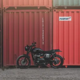 Czarny motocykl Triumph Scrambler zaparkowany na tle rdzawych kontenerów transportowych z widocznymi napisami i oznaczeniami.