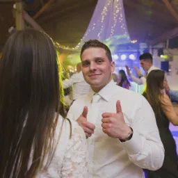 Uśmiechnięty mężczyzna w białej koszuli i krawacie, z uniesionymi kciukami do góry, podczas zabawy na parkiecie w tle z innymi gośćmi i dekoracjami świetlnymi.