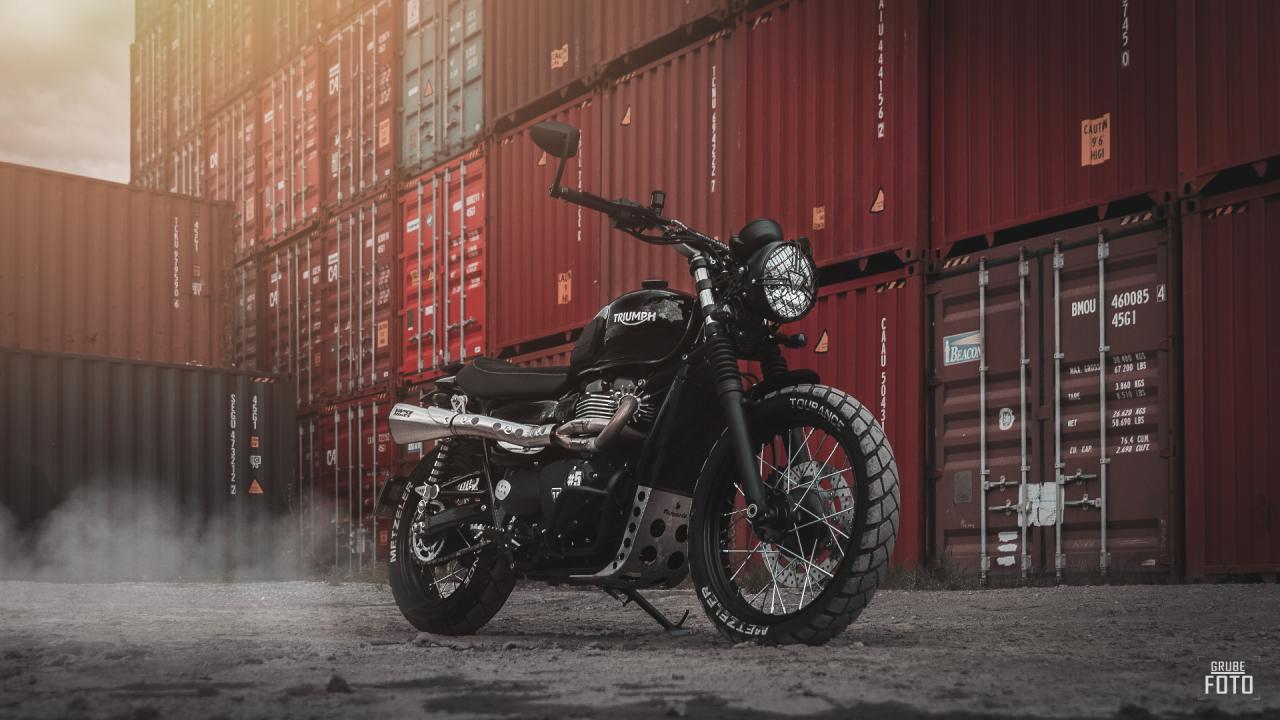 Czarny motocykl Triumph Scrambler z oponami Metzeler Tourance, zaparkowany na szarym gruncie na tle czerwonych kontenerów transportowych w porcie, z efektem dymu unoszącego się spod kół.