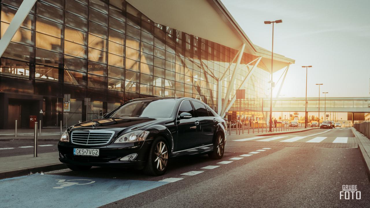 Czarna limuzyna Mercedes zaparkowana na wyznaczonym miejscu dla osób niepełnosprawnych przed nowoczesnym budynkiem lotniska w promieniach zachodzącego słońca.