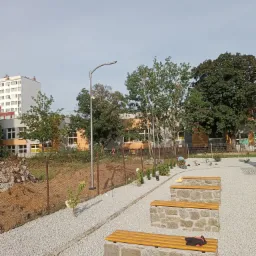 Nowo zagospodarowany teren rekreacyjny z ławkami z kamienną podstawą i drewnianym siedziskiem, ścieżka wysypana białym żwirem, w tle budynek szkoły i lampa oświetleniowa. Osoba pracuje przy...