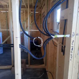 Przygotowanie instalacji alarmowej i monitoringu w domu szkieletowym: czarne karbowane rury prowadzone wewnątrz drewnianej konstrukcji ścian, z widocznymi białymi przewodami zabezpieczonymi taśmą.