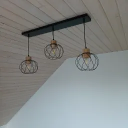 Trzy lampy wiszące z czarnymi, drucianymi kloszami i drewnianymi elementami, zamontowane na czarnej listwie, na tle drewnianego sufitu i białej ściany pod skosem.