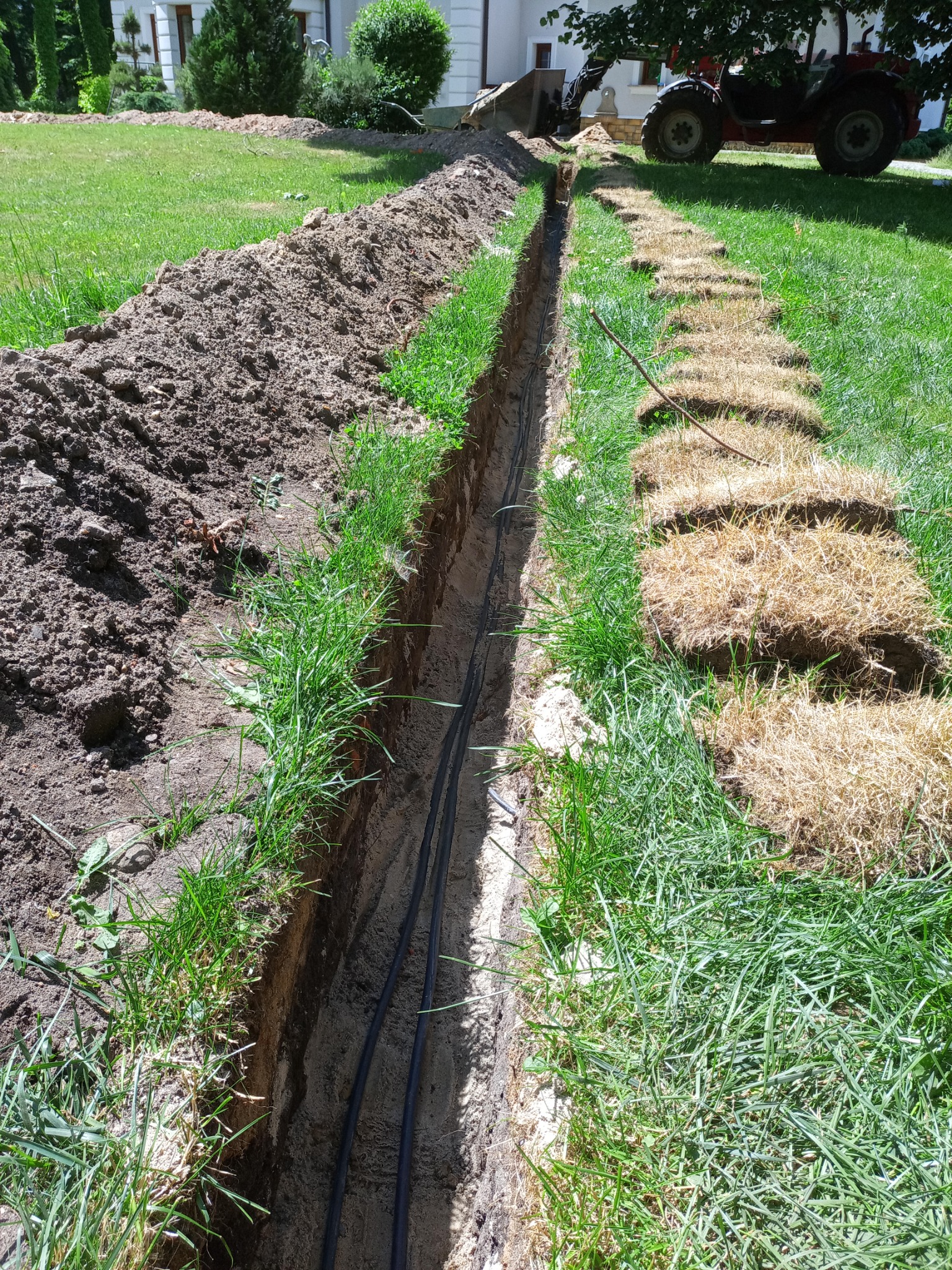 Wykopany rów z ułożonymi przewodami w Lubinie, widoczny traktor z łyżką i odłożony darń obok wykopu.