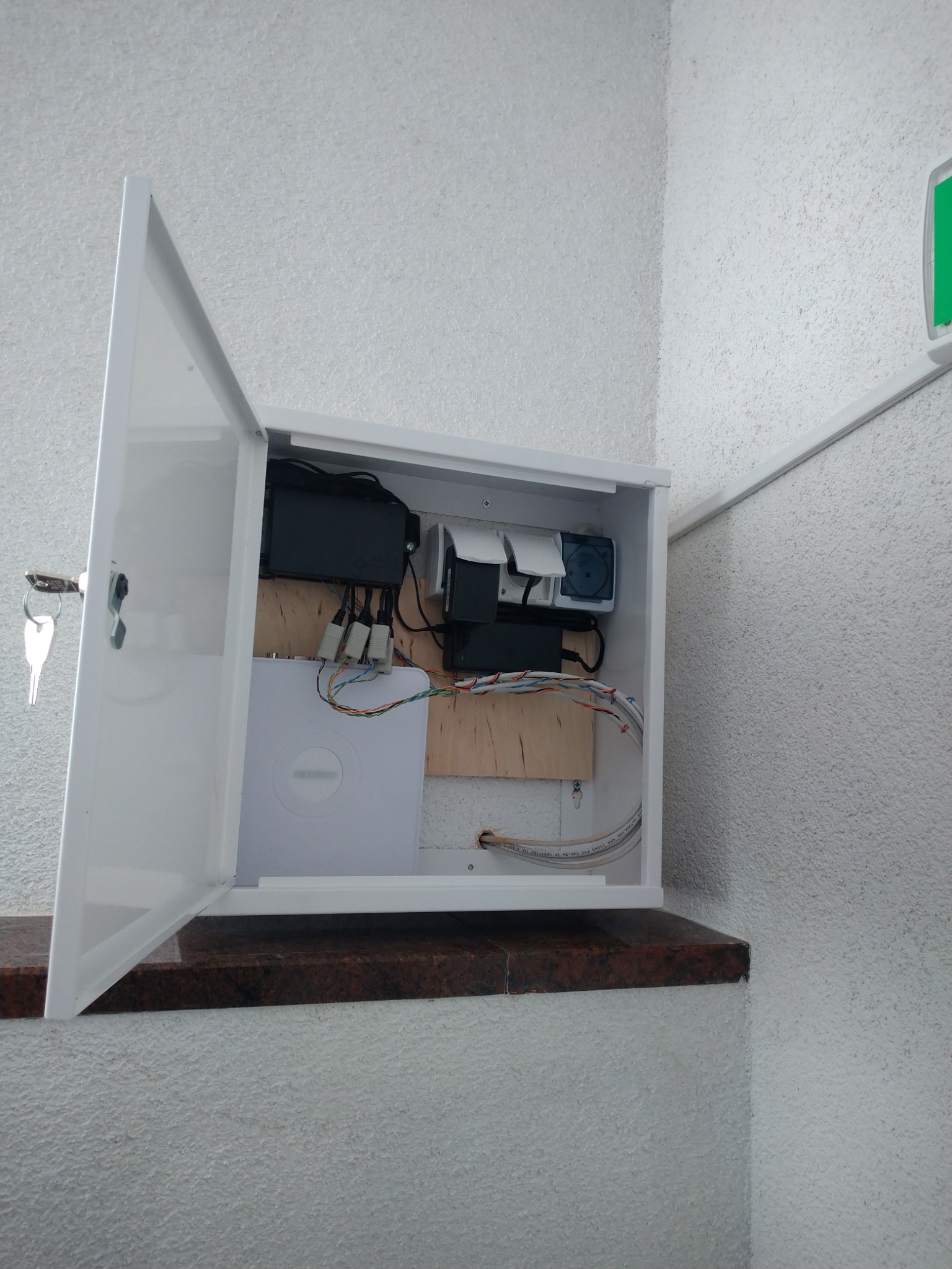 Otwarta biała szafka z zamontowanym rejestratorem monitoringu, zasilaczem i okablowaniem na drewnianej płycie, umieszczona na marmurowym parapecie przy ścianie o ziarnistej fakturze.
