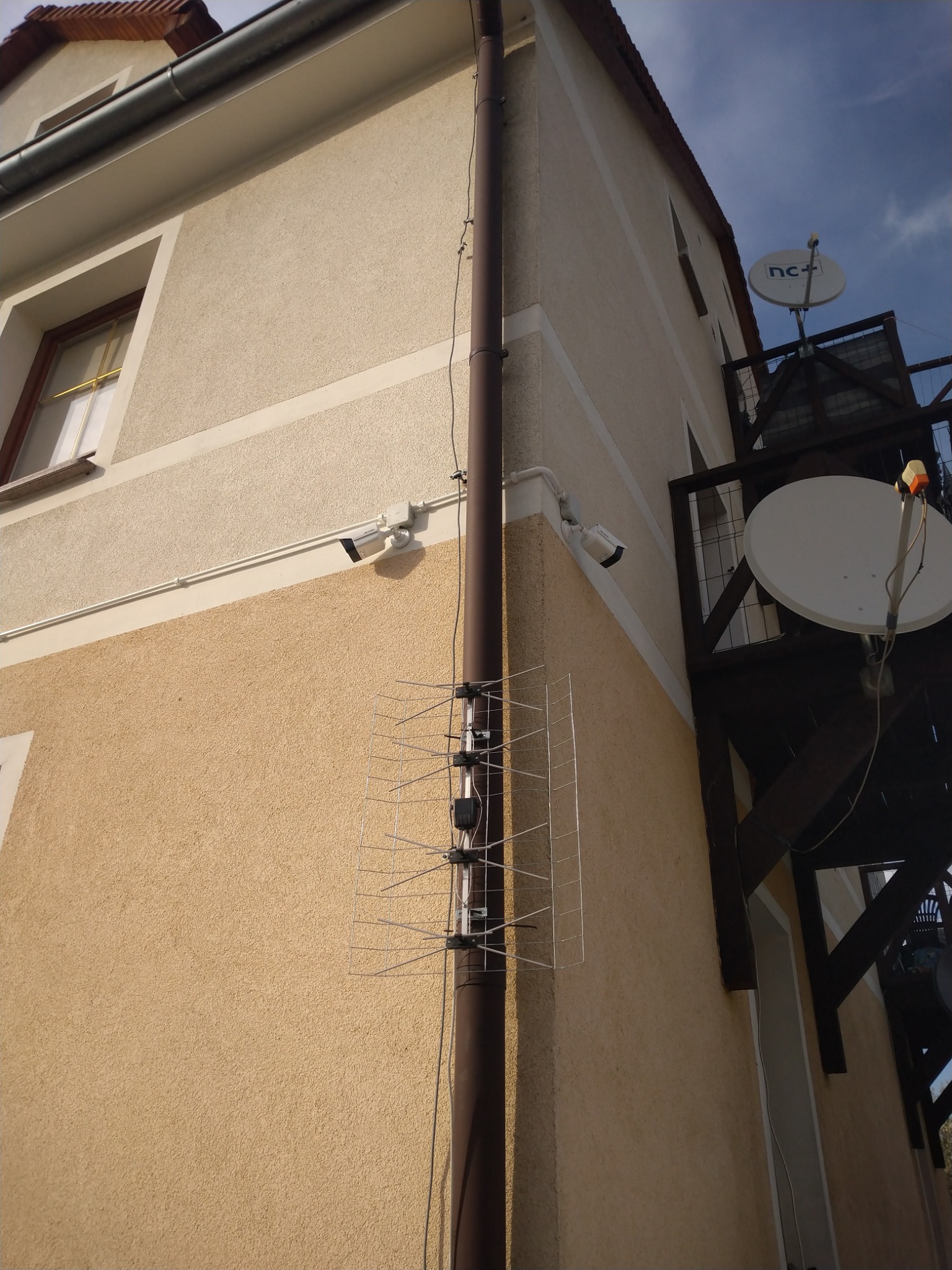 Dwie kamery monitoringu, antena telewizyjna i satelita zamontowane na elewacji dwukondygnacyjnego budynku z beżowym tynkiem.
