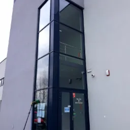 Mycie okien na wysokości w budynku biurowym z użyciem teleskopowego kija z podajnikiem wody, widoczne odbicie drzew w czystych szybach.