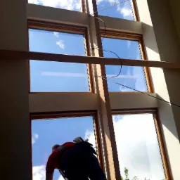 Pracownik na drabinie czyści wewnętrzną stronę wysokiego okna, z widokiem na błękitne niebo z chmurami.