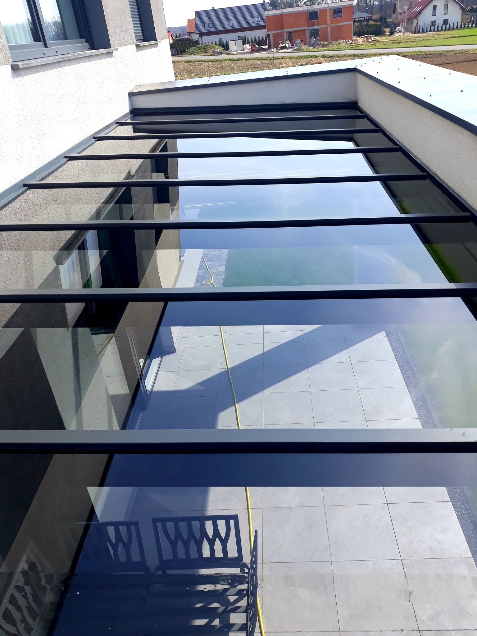 Przezroczysty szklany dach nad tarasem z widocznymi refleksami otoczenia i niebem, konstrukcja z ciemnymi ramami.