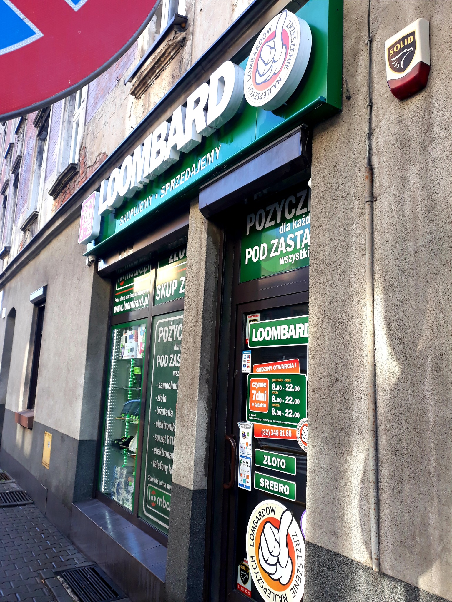 Fasada lombardu 'LOOMBARD' z widocznymi szyldami informującymi o skupie i sprzedaży, godziny otwarcia i asortyment przyjmowany pod zastaw, z naklejkami zrzeszenia lombardów na drzwiach wejściowych.