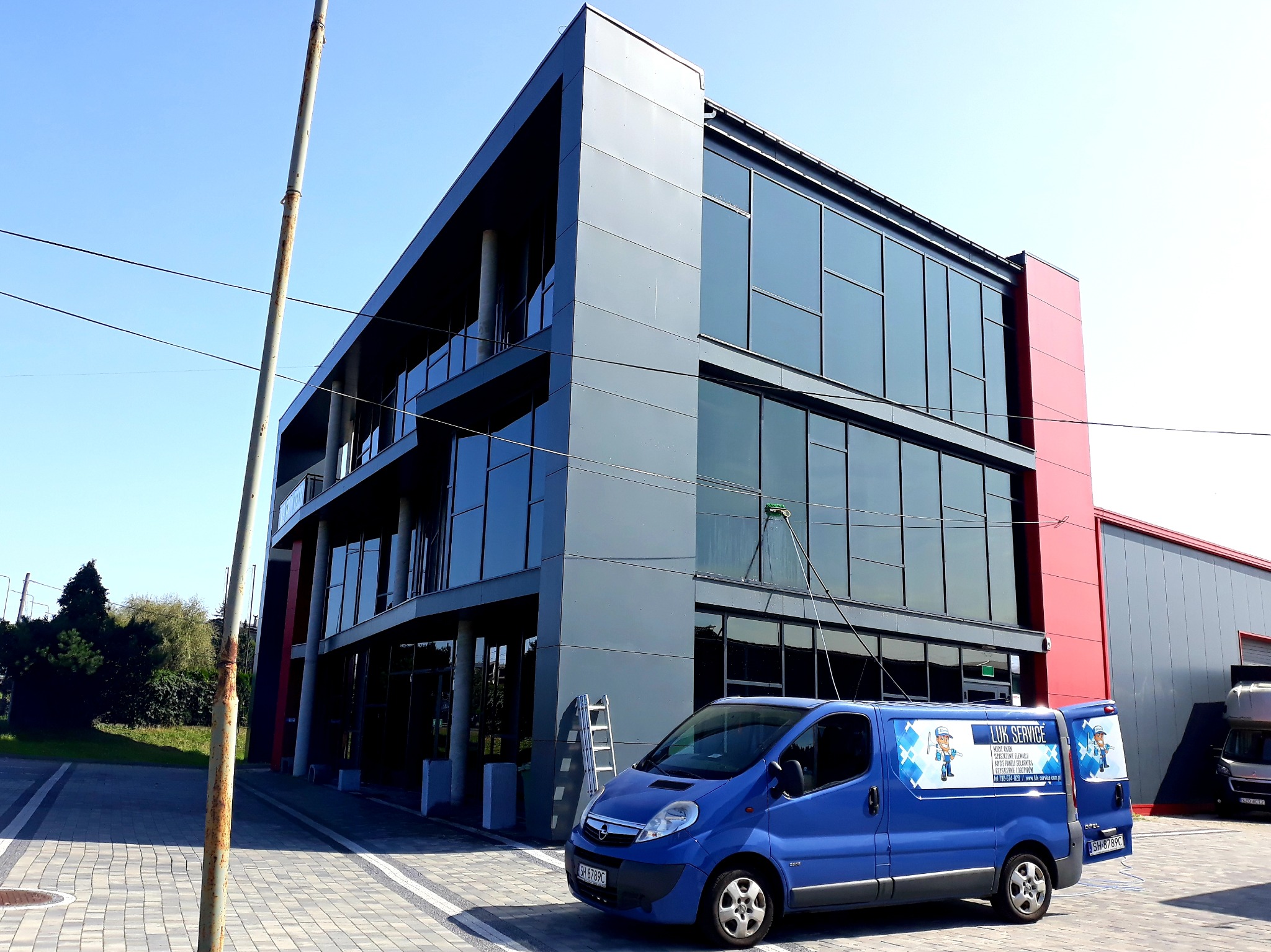 Nowoczesny budynek biurowy z szarymi panelami i dużymi oknami, czyszczony z zewnątrz za pomocą długiego drążka z myjką; na pierwszym planie niebieski samochód dostawczy z logo firmy świadczącej...