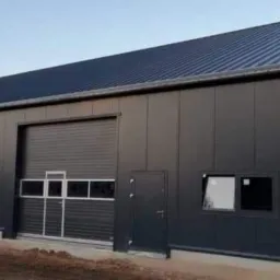 Zewnętrzny widok nowo wybudowanej hali z szarych płyt warstwowych z dwoma bramami garażowymi i drzwiami wejściowymi, widoczne rynny i fundamenty, w tle pole.