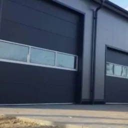 Nowoczesna hala przemysłowa z dwoma segmentowymi bramami garażowymi w kolorze grafitowym, z poziomymi oknami, widok z zewnątrz na tle błękitnego nieba.