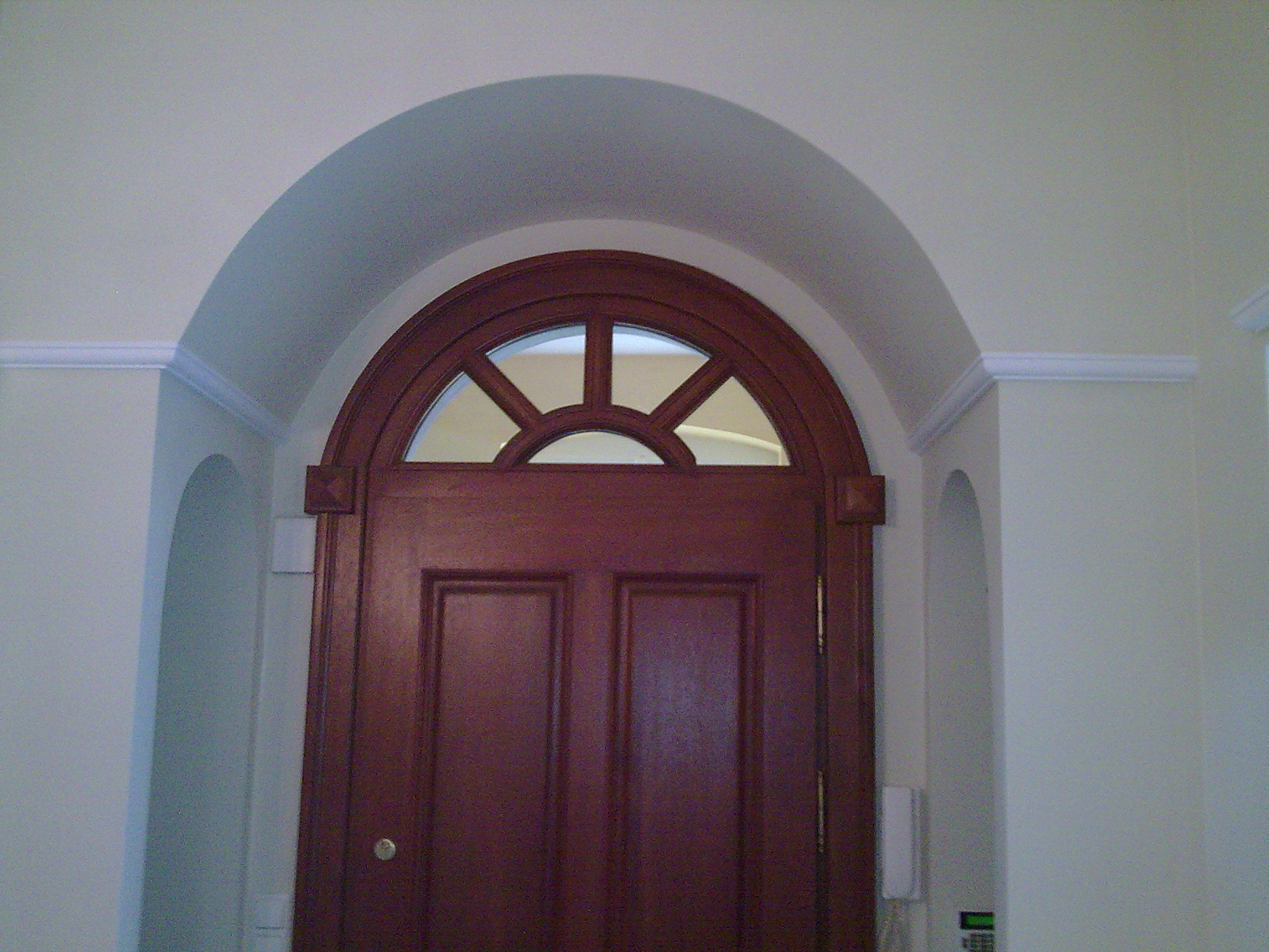 Ciemnobrązowe, dwuskrzydłowe drzwi z półokrągłym przeszkleniem w górnej części, umieszczone w łukowatym przejściu w jasnym pomieszczeniu.