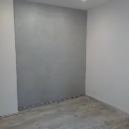 Pokój po świeżo wykonanych pracach wykończeniowych: ściana pokryta dekoracyjną szarą farbą z efektem betonu, panele podłogowe imitujące drewno, widoczne gniazdko elektryczne i listwy przypodłogowe.