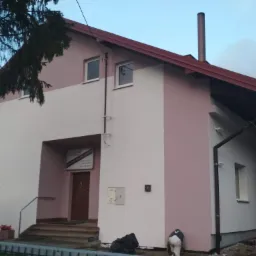 Dwukondygnacyjny budynek po remoncie elewacji, pomalowany w dwukolorowej tonacji: dolna część w odcieniach różu, górna biała. Widoczny fragment dachu z kominem, okna oraz pracownik w kombinezonie...