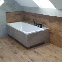 Nowoczesna łazienka na poddaszu z wanną obudowaną szarą płytką, drewnopodobną okładziną ścienną i kabiną prysznicową ze szklanymi drzwiami.