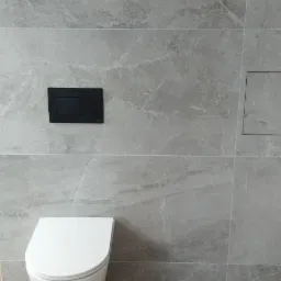 Szare płytki imitujące marmur na ścianie łazienki, z widocznym białym kompaktem WC, czarnym przyciskiem spłuczki i ukrytym inspekcyjnym włazem rewizyjnym.