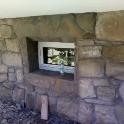 Okładzina z nieregularnego kamienia elewacyjnego wokół okna, widoczna butelka wody na parapecie i resztki gruzu u podnóża ściany w Szymbarku.