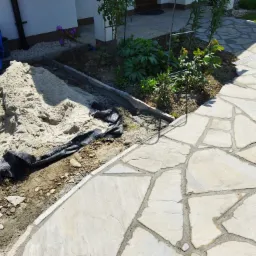 Fragment tarasu z kamienia naturalnego w trakcie budowy, widoczny piasek, krawężnik i fragment ogrodu z roślinami.