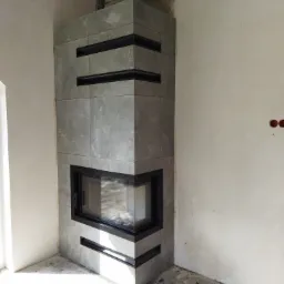 Obudowa kominka z szarych płytek imitujących kamień, z czarnymi wstawkami, w rogu pomieszczenia w trakcie remontu, widoczne niedoskonałości ścian i podłogi.