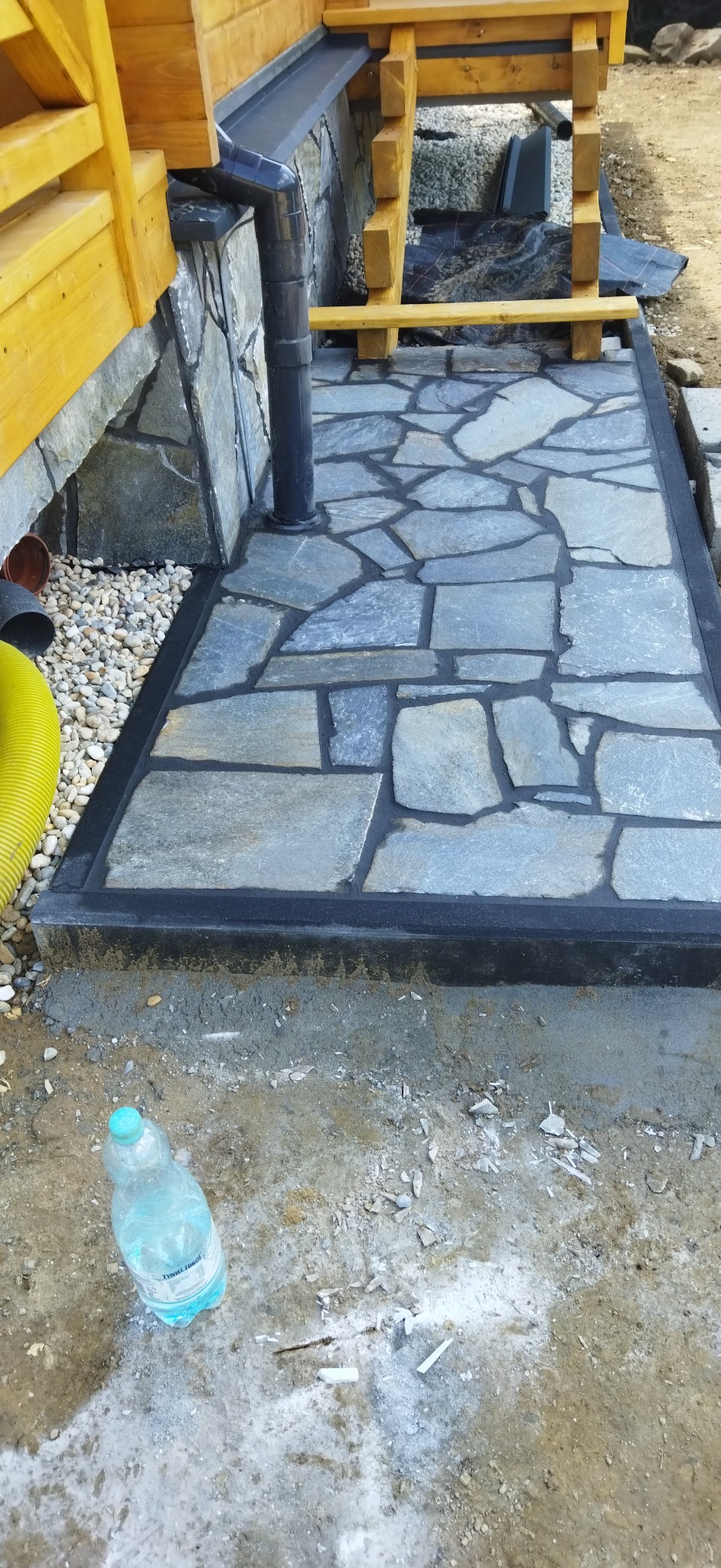 Fragment tarasu z nieregularnych płyt kamiennych w odcieniach szarości, obramowany czarną listwą, z widoczną rurą spustową i fragmentem drewnianej konstrukcji budynku w tle, oraz butelką wody...