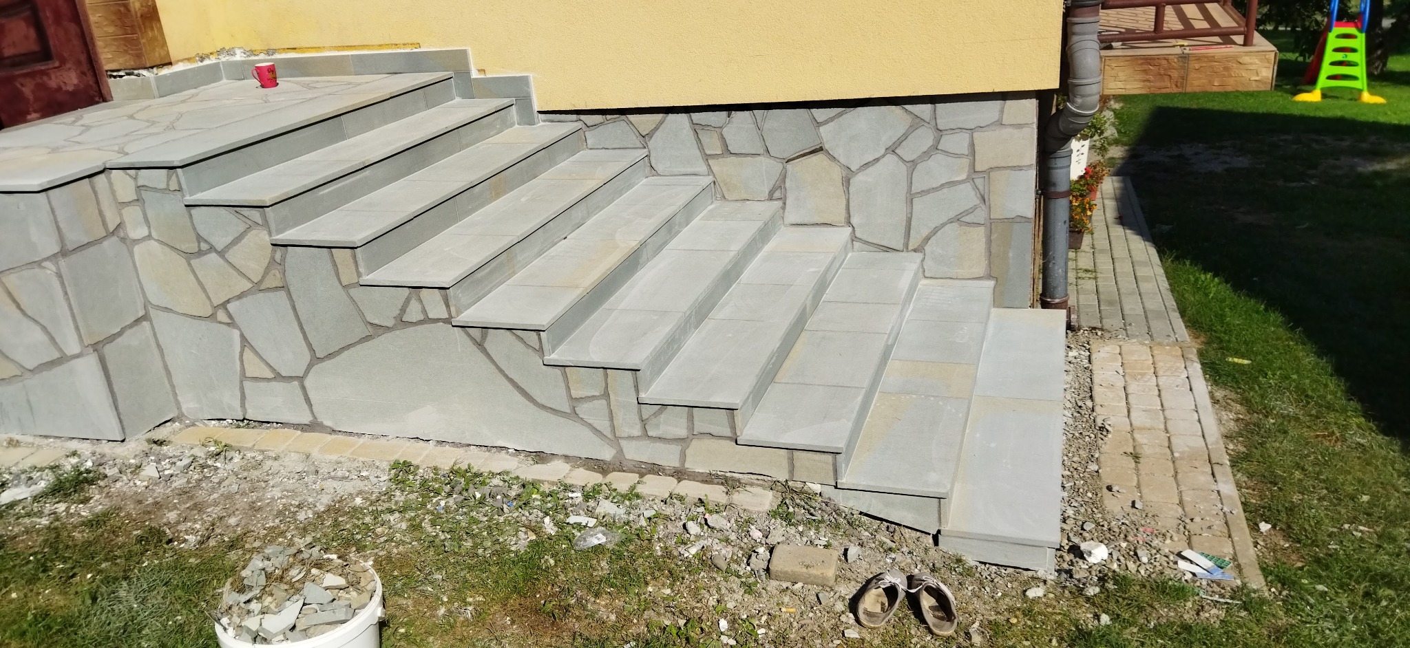 Kamienne schody wejściowe do domu z szarych płyt, ściana obłożona nieregularnym kamieniem, kubek na pierwszym stopniu, buty robocze obok schodów.