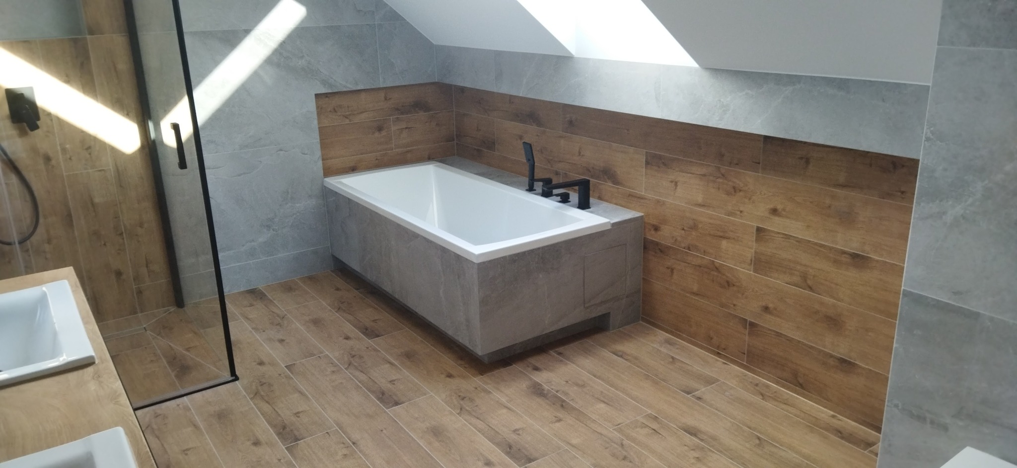 Nowoczesna łazienka na poddaszu z wanną obudowaną szarą płytką, drewnopodobną okładziną ścienną i kabiną prysznicową ze szklanymi drzwiami.