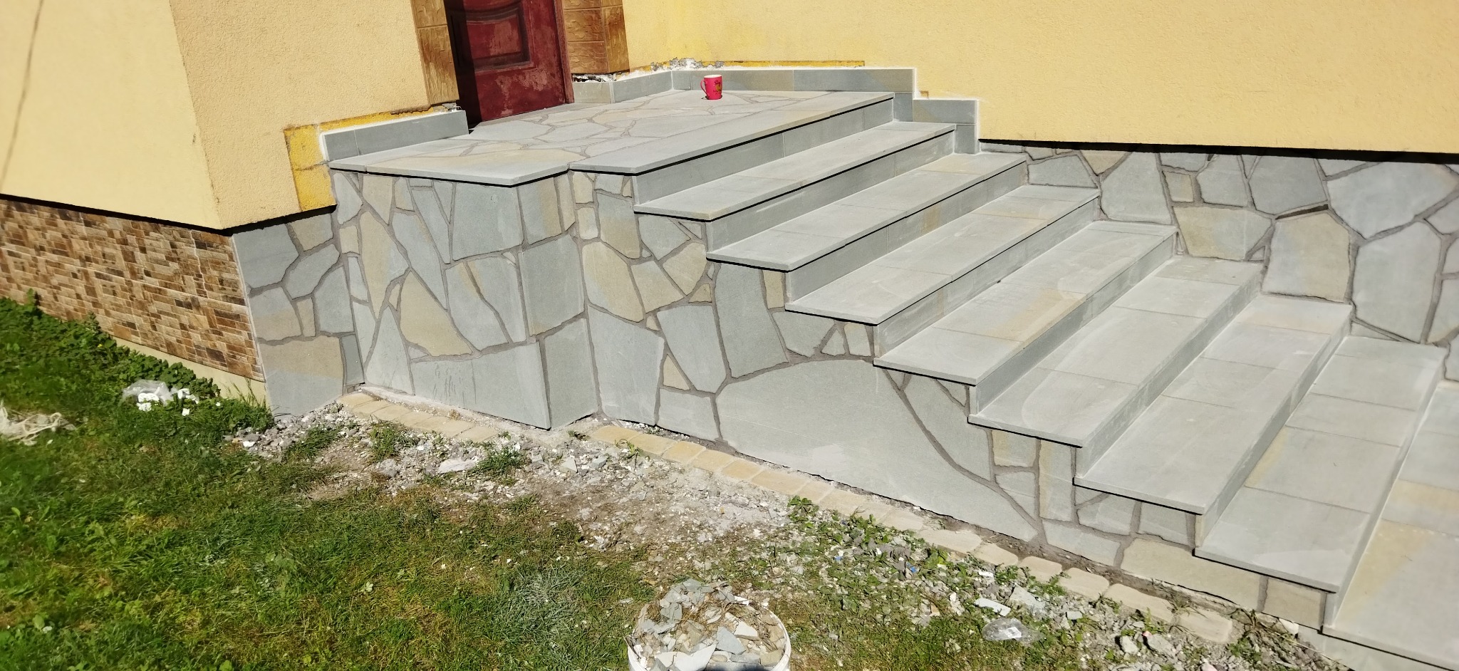 Kamienne schody wejściowe do domu, wykończone nieregularnym kamieniem elewacyjnym, z czerwoną filiżanką na górnym podeście.