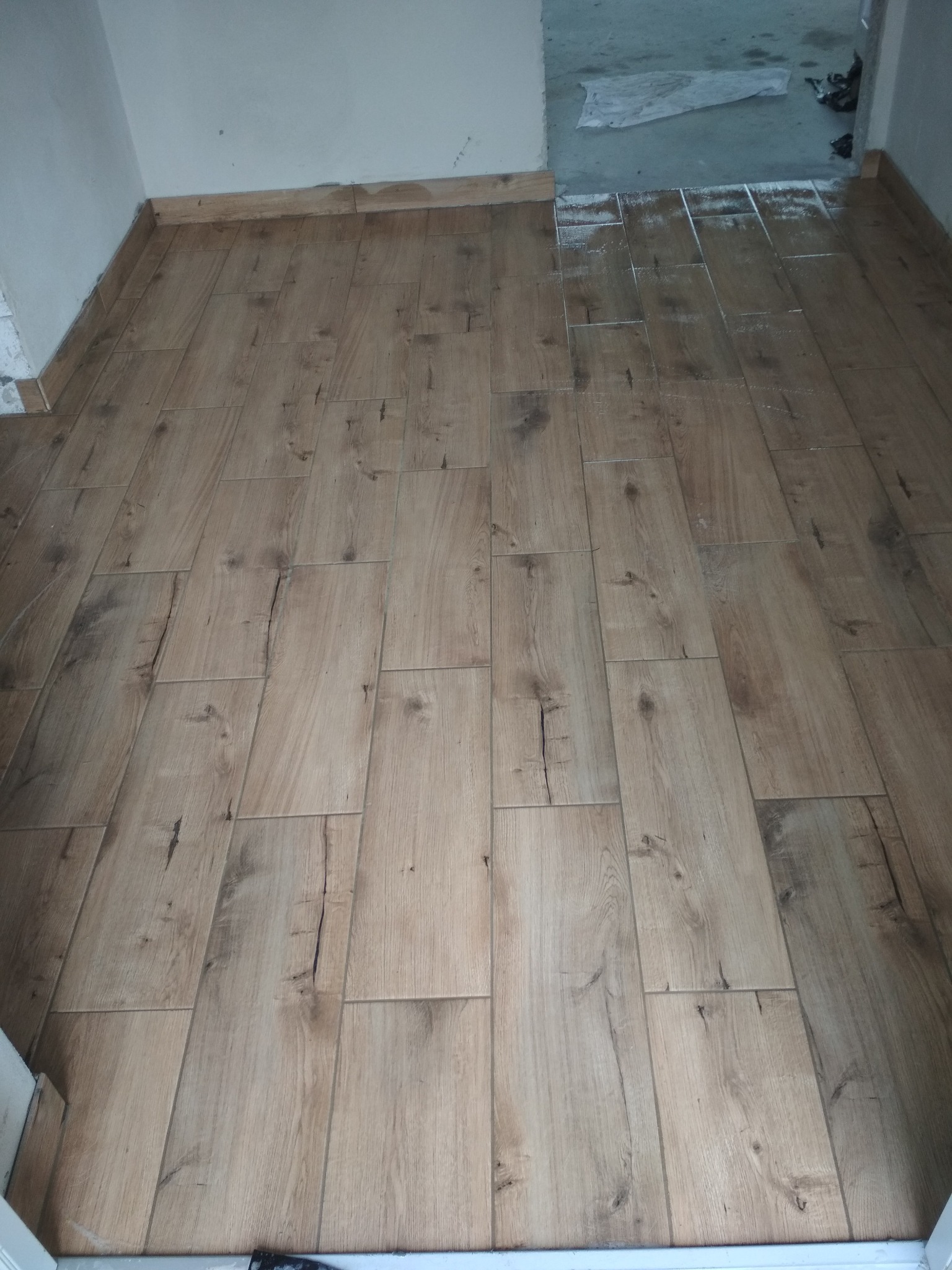 Ułożona podłoga z płytek imitujących drewno, widoczne fugi i listwy przypodłogowe, w tle niedokończone pomieszczenie z resztkami zaprawy.