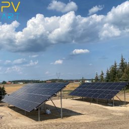 PV-Art - Dwa panele słoneczne na metalowych konstrukcjach, ustawione na piaszczystym terenie z widokiem na wieś i błękitne niebo z białymi chmurami.