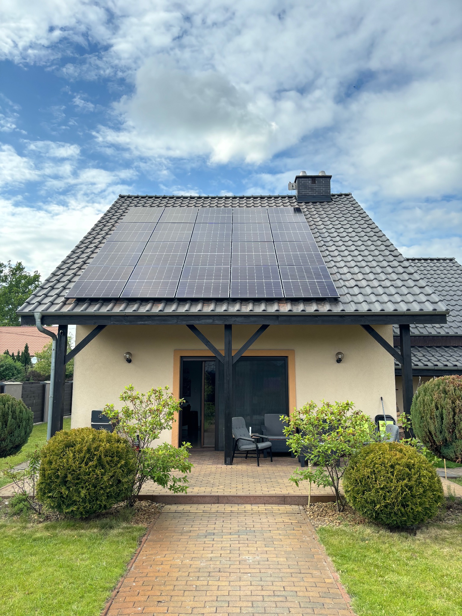 Dom jednorodzinny z panelami fotowoltaicznymi na dachu pokrytym szarą dachówką, widok z przodu na zadbany ogród z kostką brukową prowadzącą do wejścia.