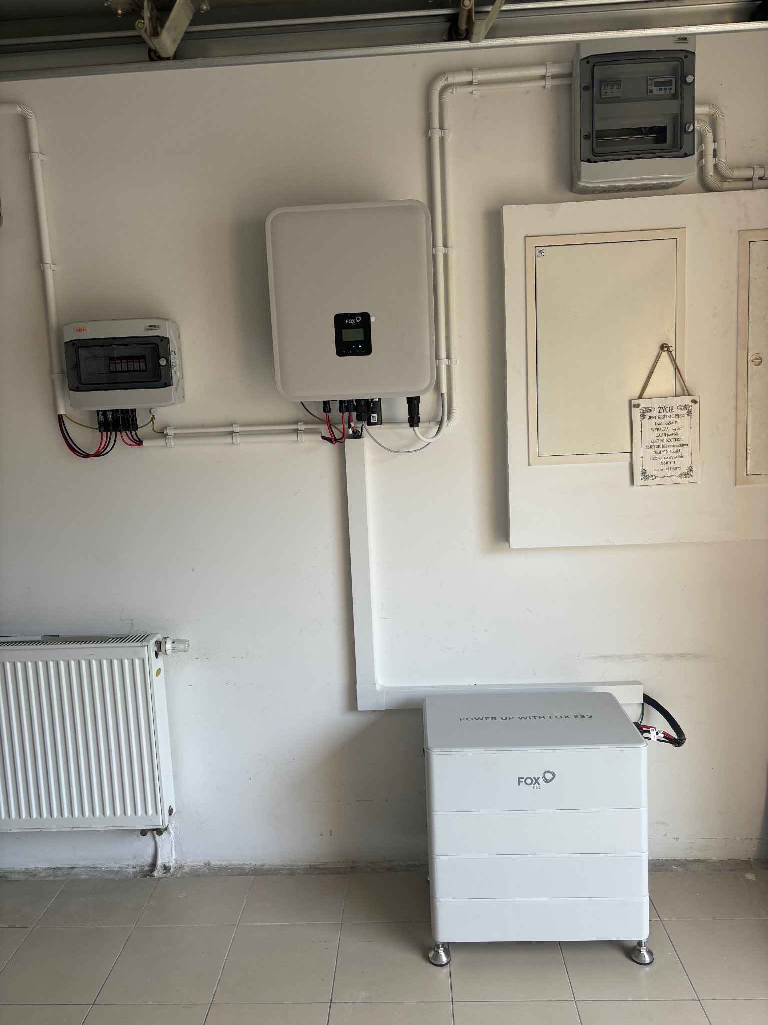 Instalacja domowa z białym inwerterem, rozdzielnicą elektryczną i modułem magazynu energii Fox ESS na tle białej ściany z widocznymi rurami instalacyjnymi i grzejnikiem.