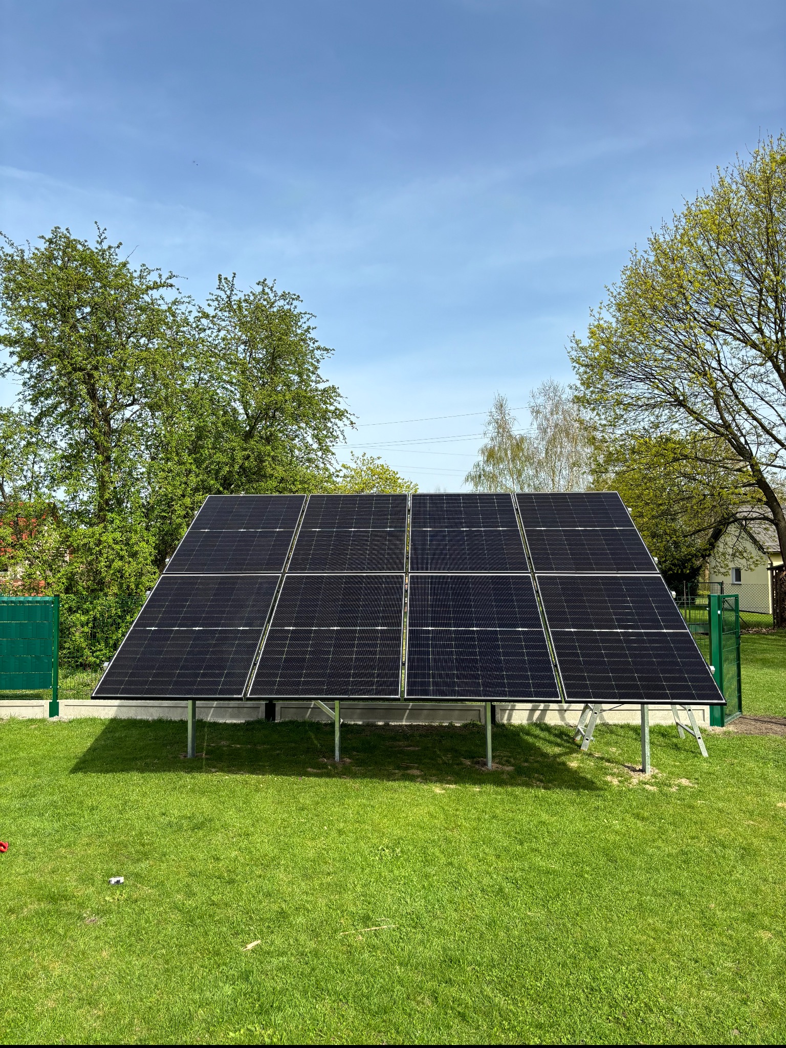 Panel fotowoltaiczny na metalowej konstrukcji, ustawiony na trawniku w ogrodzie, widoczny zielony płot i drzewa w tle, błękitne niebo.