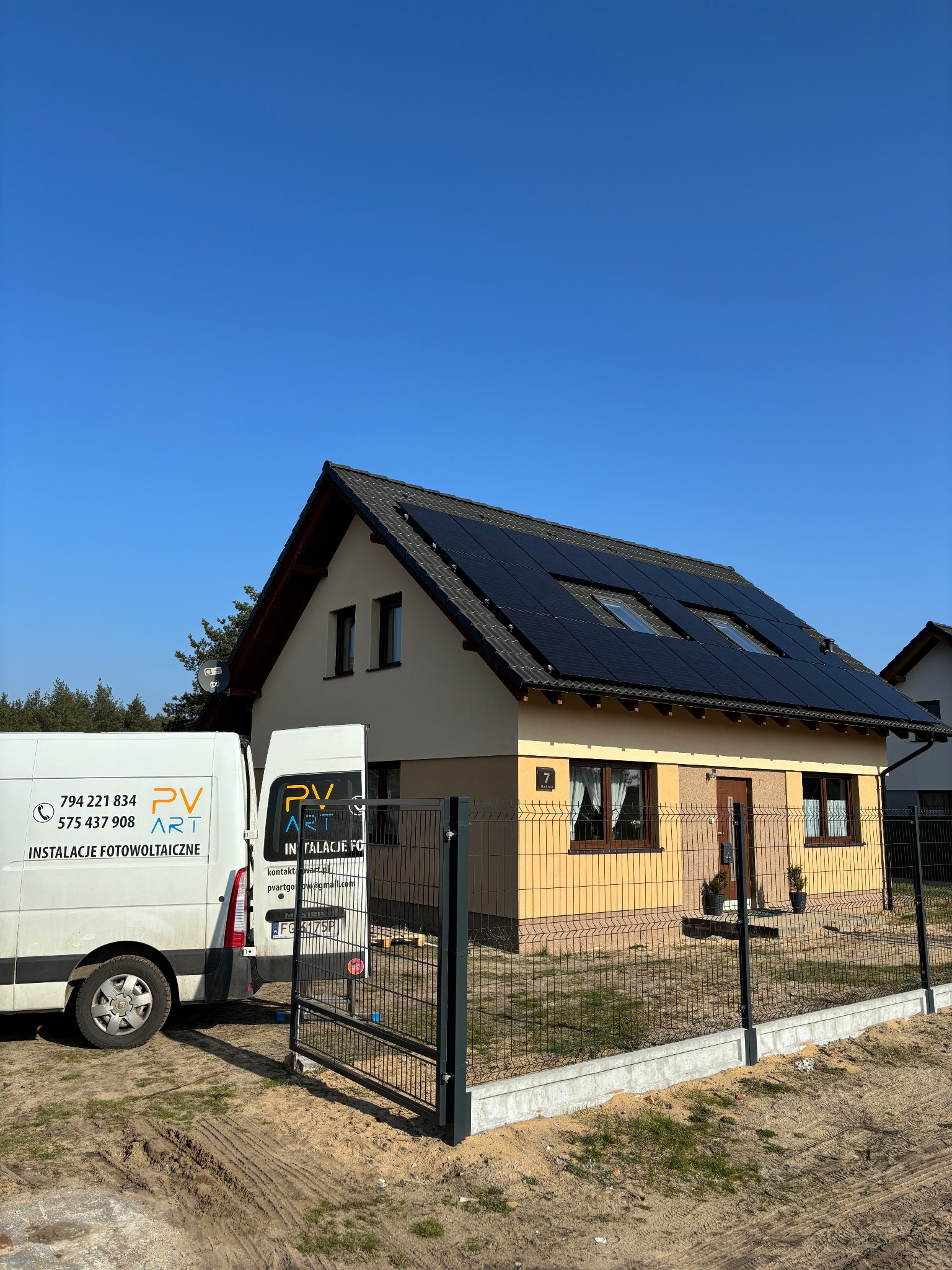Dom jednorodzinny z zamontowanymi panelami słonecznymi na dachu, widoczny samochód dostawczy z logo firmy instalującej panele, ogrodzenie posesji