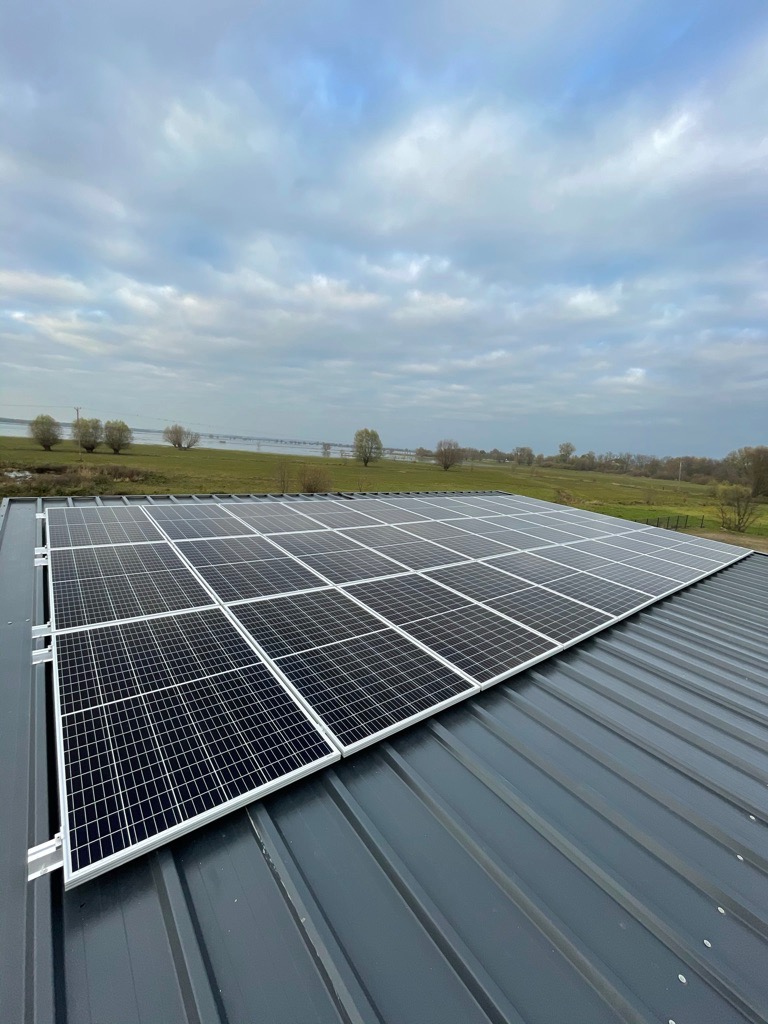 Panele słoneczne zamontowane na szarym, metalowym dachu, z widokiem na zielone pole i pochmurne niebo w tle.