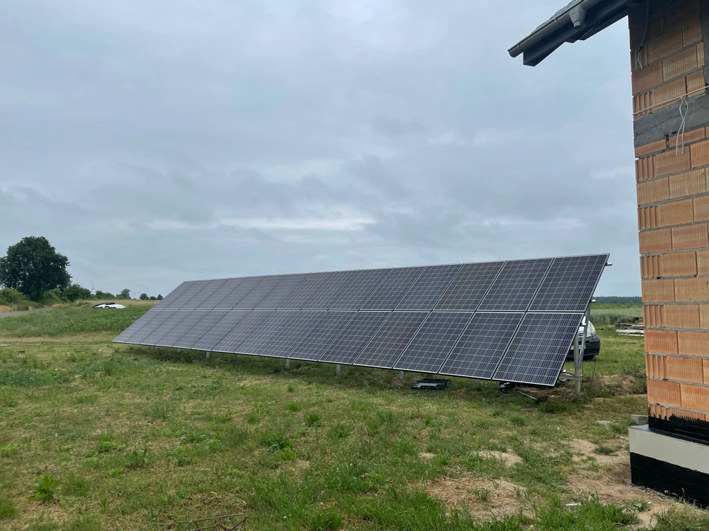 Rząd paneli słonecznych na metalowych podporach, zainstalowany na trawiastym terenie obok budynku w trakcie budowy, widoczny samochód w tle.