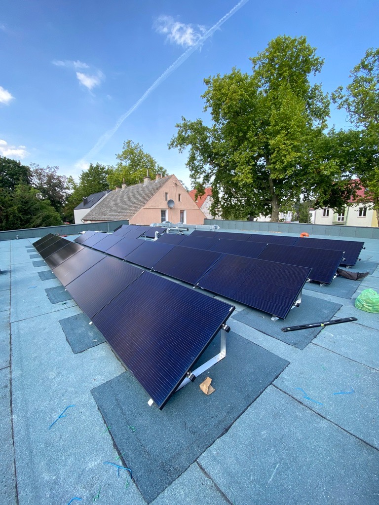 Panele fotowoltaiczne w trakcie montażu na płaskim dachu pokrytym papą, widoczne narzędzia i elementy montażowe, w tle budynek mieszkalny i drzewa.