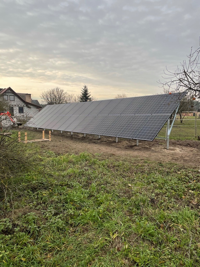 Panel słoneczny na metalowej konstrukcji w wiejskim otoczeniu, z domem w tle i pochmurnym niebem.