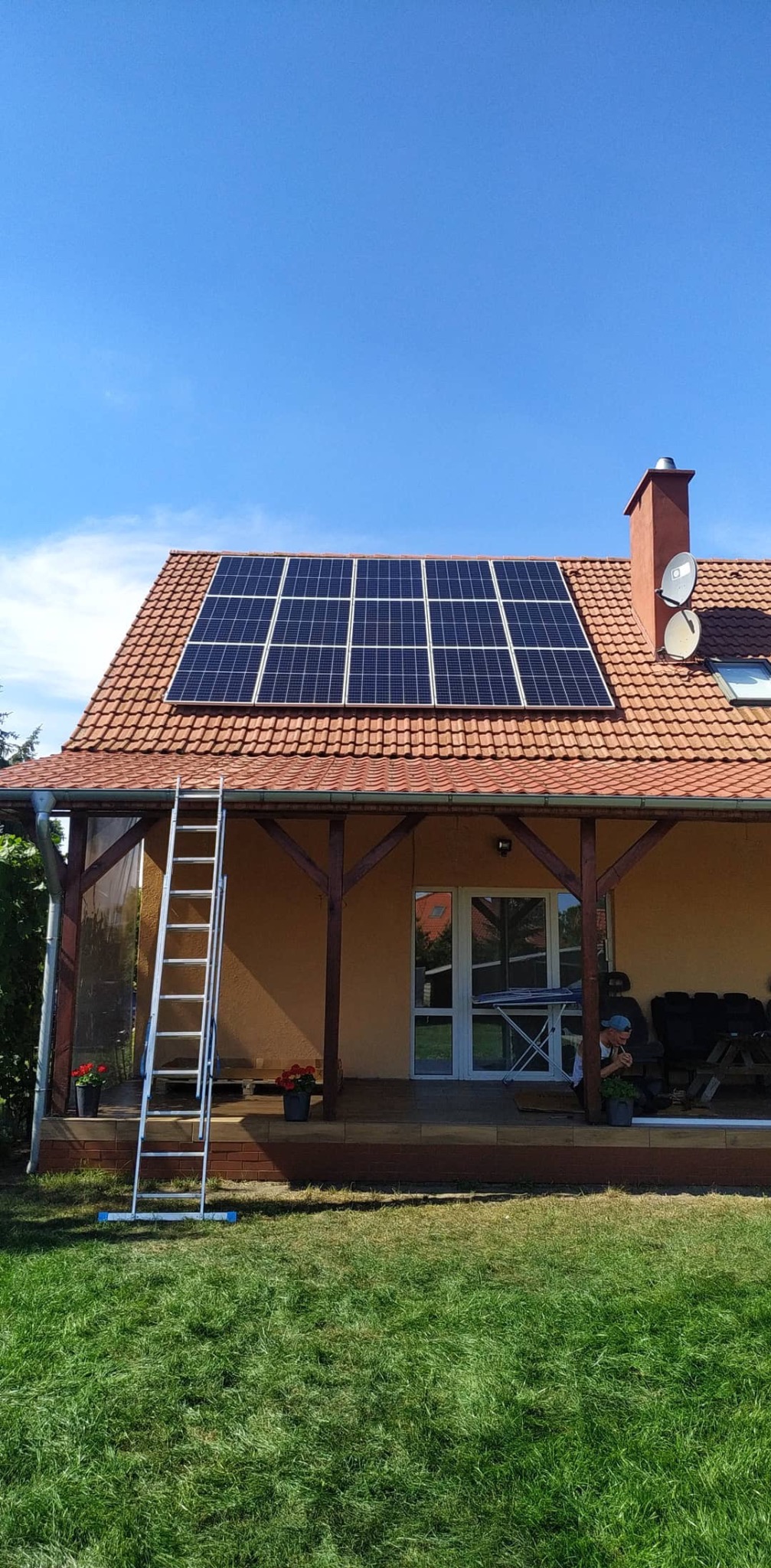 Panele słoneczne na dachu domu jednorodzinnego z czerwoną dachówką, drabina oparta o dach, błękitne niebo w tle.