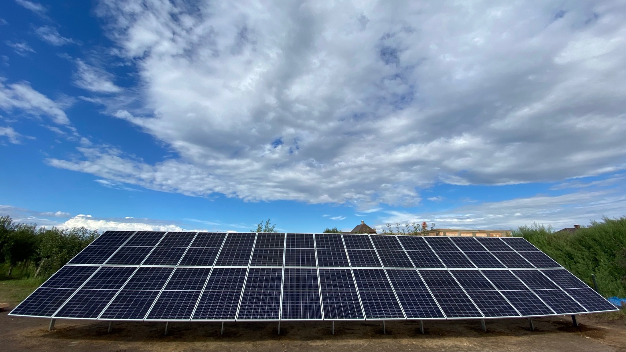 Panel słoneczny na metalowych wspornikach, widoczny na tle błękitnego nieba z białymi chmurami, otoczony zielenią krzewów i drzew