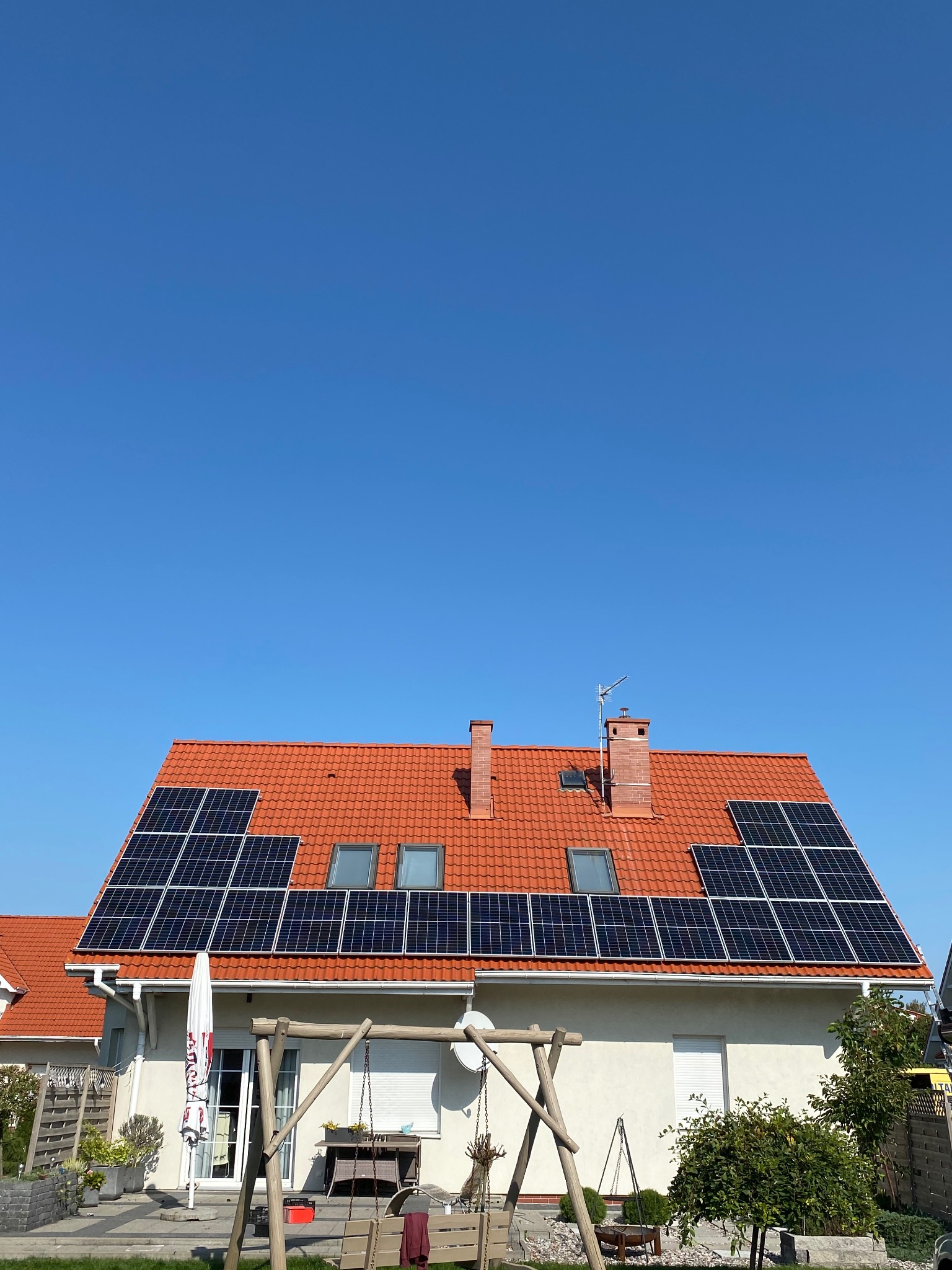 Dom jednorodzinny z czerwoną dachówką, na której zamontowane są panele słoneczne; widoczny fragment ogrodu z huśtawką, jasne, bezchmurne niebo.