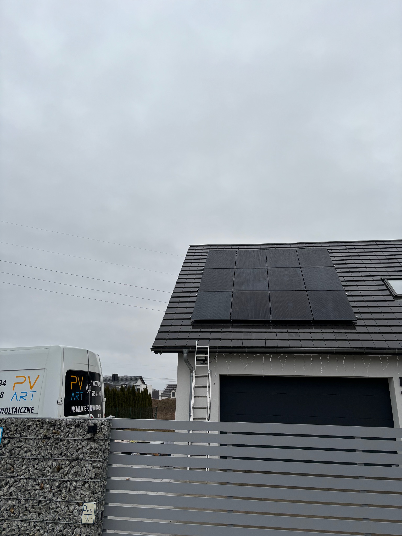 Panele słoneczne zamontowane na dachu domu jednorodzinnego z szarą dachówką, widoczna drabina oparta o ścianę, w tle zaparkowany biały bus z logo firmy.