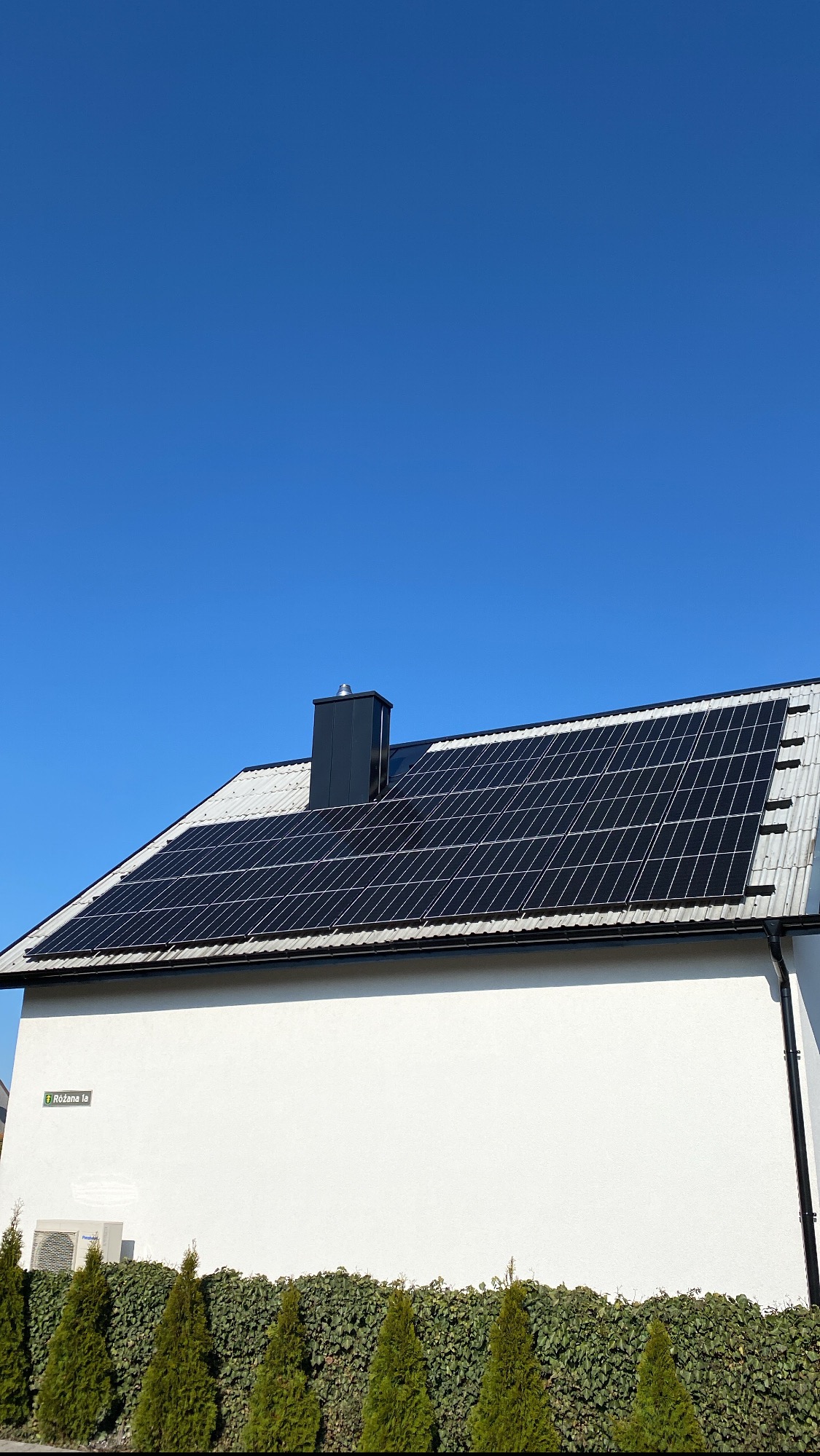 Panele słoneczne na dachu domu jednorodzinnego z czarnym kominem, widok z dołu na tle błękitnego nieba, żywopłot z iglaków wzdłuż ściany.