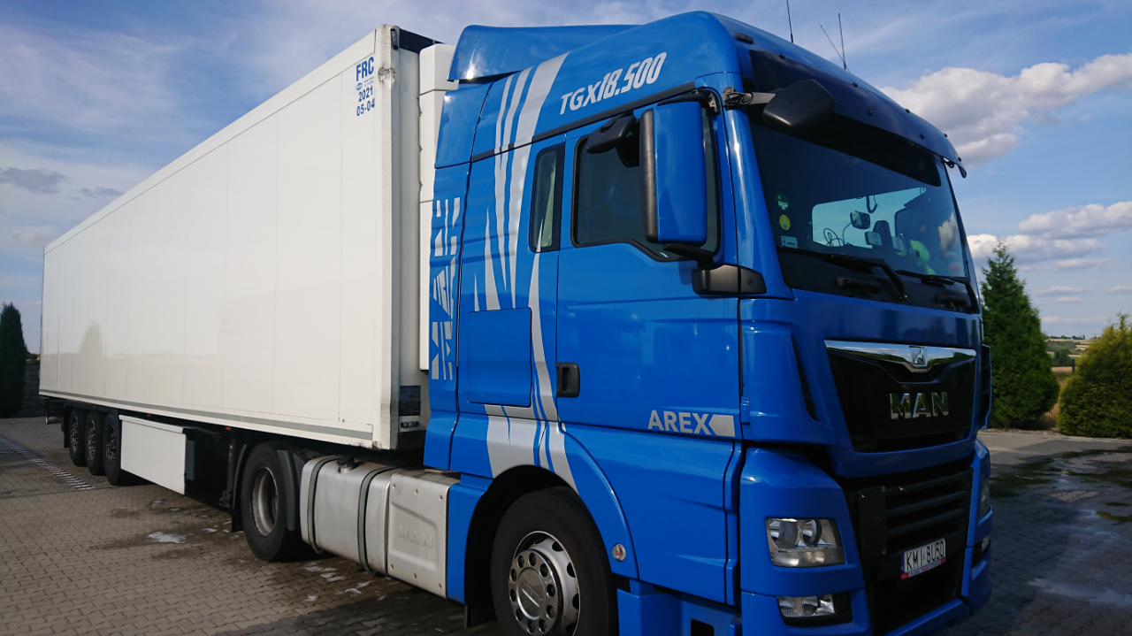 Niebieska ciężarówka MAN TGX 18.500 z białą naczepą chłodniczą, zaparkowana na brukowanym placu w słoneczny dzień.