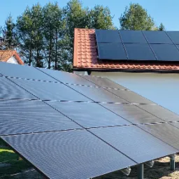 Instalacja fotowoltaiczna na gruncie z widocznymi panelami skierowanymi ku słońcu, w tle dom z panelami na dachu i drzewami.
