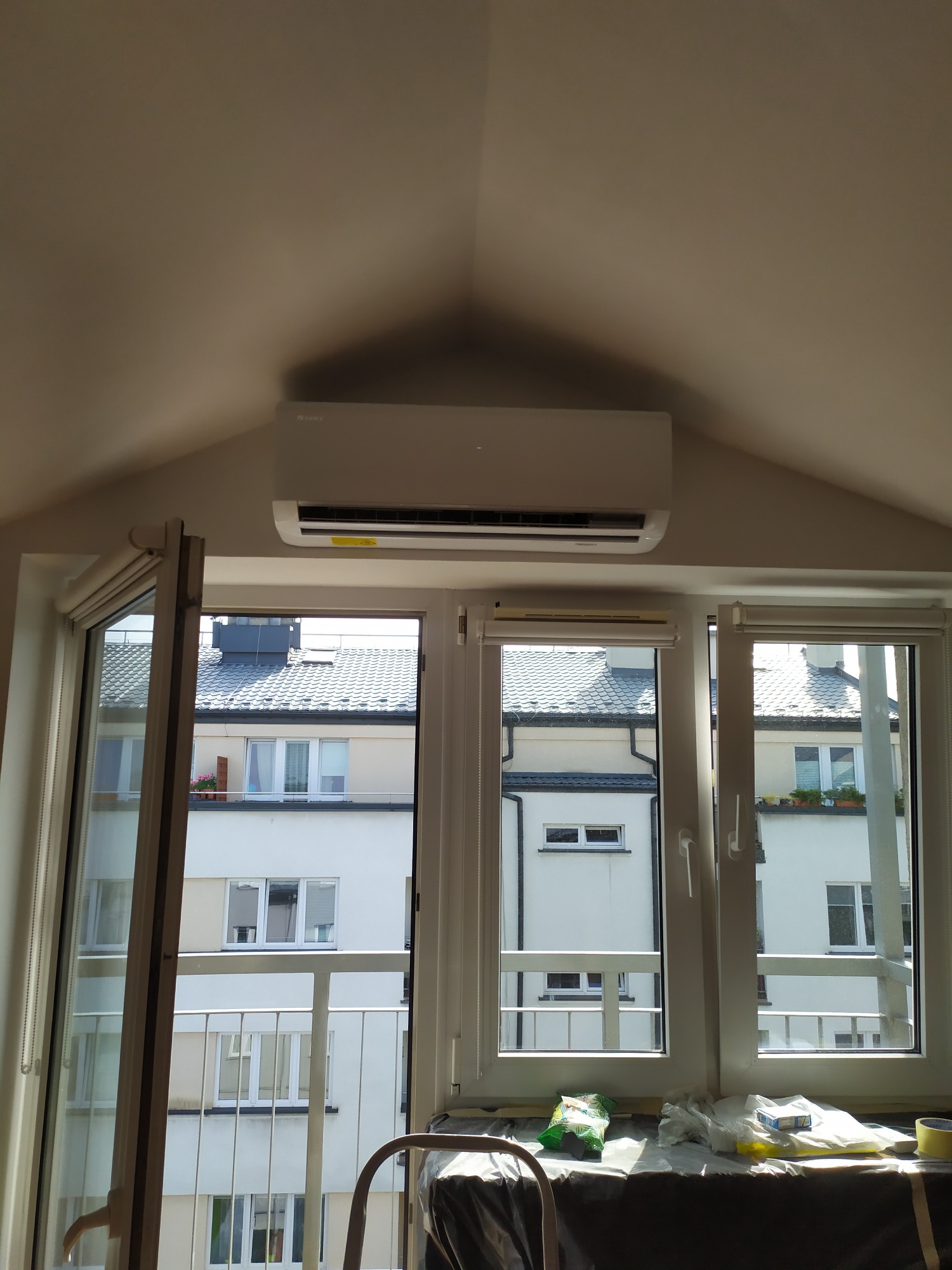 Biały klimatyzator Gree zamontowany nad oknem w pokoju na poddaszu z widokiem na budynki mieszkalne, zabezpieczony parapet przed zabrudzeniem podczas montażu.
