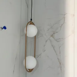 Lampa wisząca z dwoma białymi kulami w złotej, podłużnej ramie na tle ściany wyłożonej płytkami imitującymi marmur.